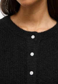 Cardigan en tricot noir avec un col rond, trois fermetures à boutons blancs et un tissu texturé au toucher doux.