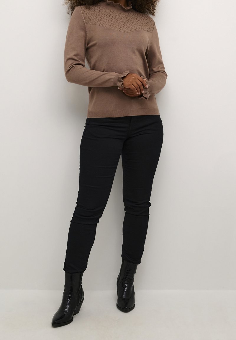 Ruskea neule, jossa on teksturoitu olkapään yksityiskohta, pitkät hihat ja rypytyshelma, yhdistettynä mustiin istuviin housuihin ja mustiin nilkkureihin.