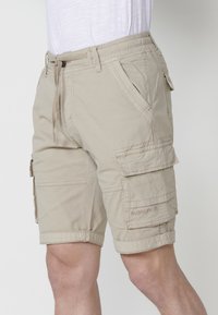 Shorts cargo bej cu talie elastică și șnur. Dispune de buzunare laterale și buzunare cargo pe ambele coapse, fabricate dintr-un material durabil.