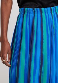 Spódnica plisowana w odcieniach niebieskiego i zielonego. Elastyczny pas z pionowymi paskami i miękką fakturą. Ręka widoczna z pierścionkiem.