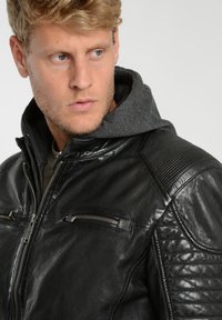 Sort læderjakke med lynlås foran, grå hætte, ribbede skulderdetaljer og to lynlåslommer på brystet. Glat tekstur og tætsiddende pasform.