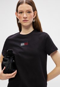 Czarny bawełniany T-shirt z małym, wielokolorowym logo z odrobiną kryształków. Model trzyma w ręce teksturowany czarny futrzany worek.