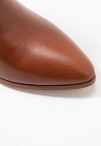 Chaussure en cuir marron à bout pointu, présentant une texture lisse et une semelle fine avec un détail de couture subtil près de la base.