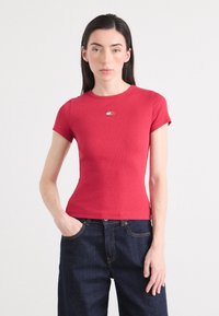 Modèle portant un t-shirt rouge ajusté avec un petit logo sur la poitrine et un jean en denim foncé taille haute, se tenant contre un fond clair.