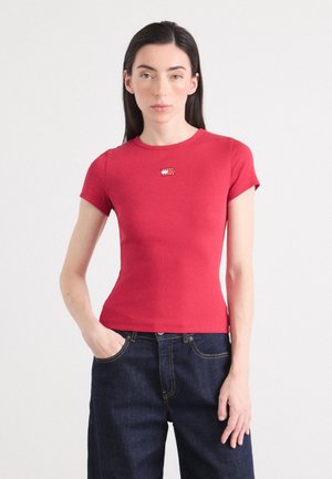 Modèle portant un t-shirt rouge ajusté avec un petit logo sur la poitrine et un jean en denim foncé taille haute, se tenant contre un fond clair.
