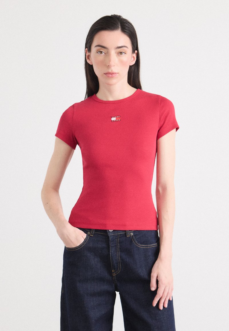 Modèle portant un t-shirt rouge ajusté avec un petit logo sur la poitrine et un jean en denim foncé taille haute, se tenant contre un fond clair.