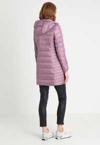 Lang, letvæg pufferjakke i lys lilla, med hætte og lodrette syninger. Sammen med sorte leggings og sølvfarvede træningssko.