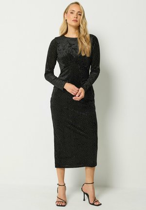 Donna con capelli lunghi biondi indossa un vestito midi nero a maniche lunghe con piccoli pois bianchi e tacchi alti neri con cinturini.