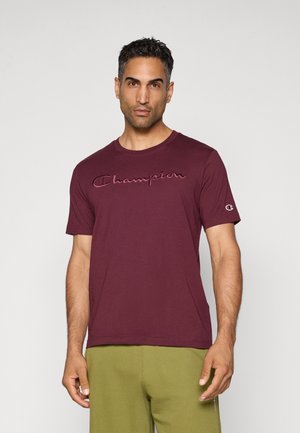 Man standing against light background wearing a maroon Champion t-shirt and olive green pants, looking forward with a neutral expression.