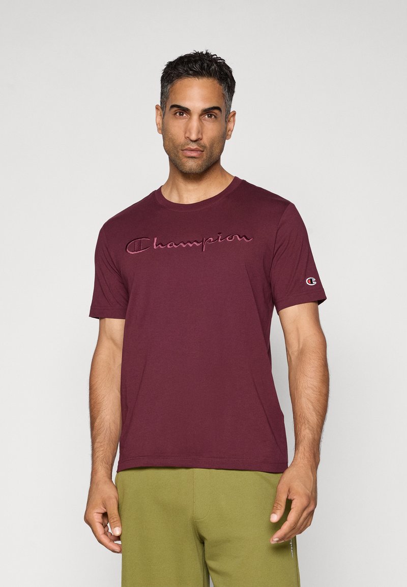 Mann, der vor einem hellen Hintergrund steht, trägt ein kastanienbraunes Champion T-Shirt und olivgrüne Hosen, sieht mit neutralem Ausdruck nach vorne.
