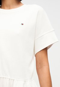 Λευκό φόρεμα με λείο πλεκτό επάνω μέρος και φαρδιά μανίκια, διακοσμημένο με ένα μικρό κεντημένο λογότυπο. Το κάτω μέρος διαθέτει σούρα φούστα με απαλή υφή.