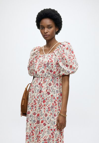 Femme portant une robe fleurie à manches bouffantes avec des fleurs rouges et bleues, portant un sac à bandoulière marron et un bracelet en or.
