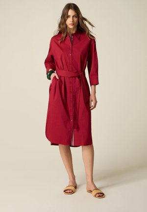 Kobieta ubrana w czerwoną sukienkę z guzikami sięgającą kolan, z rękawami do ćwierć łokcia, przewiązanym paskiem w talii oraz beżowymi plecionymi sandałami wsuwanymi.