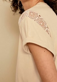 T-shirt beige avec des manches retroussées et des détails en dentelle sur l'épaule. Matériau en coton doux avec une texture lisse.