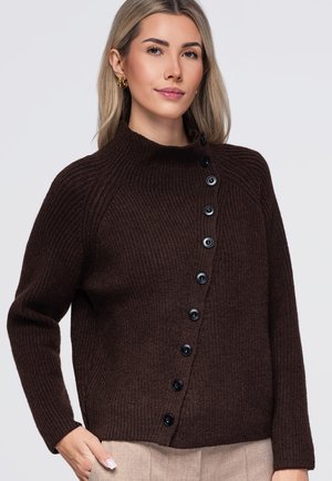 Femme aux cheveux clairs portant un pull côtelé marron foncé boutonné en diagonale et un pantalon beige, debout avec une main dans la poche.