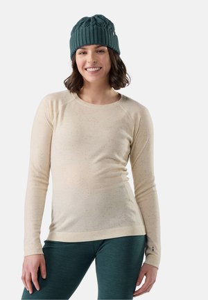 Mujer sonriente que lleva un gorro de punto verde, una camisa de manga larga beige y pantalones verde oscuro, de pie frente a un fondo claro y sencillo.