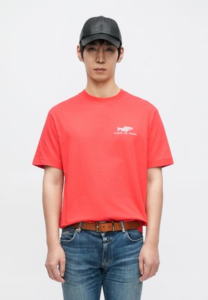 Red cotton t-shirt with short sleeves, featuring a small white graphic text design. Worn with blue jeans and a brown belt. Black cap on head.