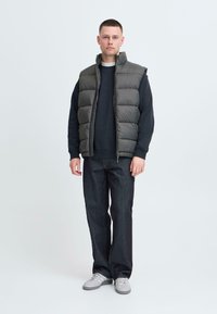 Dunkelgraue Weste mit Steppdesign, mit Reißverschluss vorne, kombiniert mit einem schwarzen Pullover und dunklen Jeans. Die Schuhe sind hellgraue Sneakers.