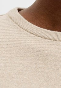 Colletto di maglione beige con lavorazione a maglia strutturata. Il tessuto è morbido con un motivo sottile e fine e un dettaglio a coste sul collo, che mostra una finitura liscia.