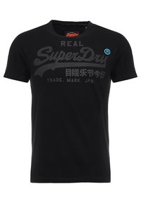 Camiseta de algodón negra con cuello redondo, que presenta un logo gris "SuperDry" texturizado y un acento azul. Diseño simple, ajustado y sin gráficos.