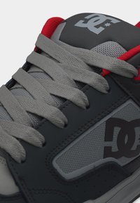 Γκρι και μαύρο sneaker DC με κόκκινη επένδυση, χοντρές γκρι κορδόνια, πάνελ από πλέγμα και λογότυπο DC στη γλώσσα και στην πλαϊνή πλευρά.