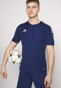 Camisa atlética de color marino con mangas cortas, escote en V y tres rayas blancas en los hombros. Sosteniendo un balón de fútbol blanco con acentos de estrellas coloridas.
