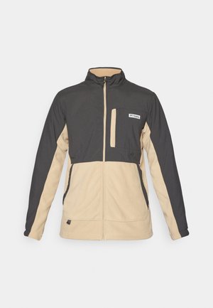 Fleecejakke med lynlås foran, sorte og beige farveblokke, høj krave og en brystlomme med et logo. Blød tekstur og afslappet design.