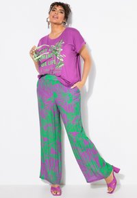 T-shirt XXL viola con testo grafico verde, abbinato a pantaloni a gamba larga con motivo floreale verde su sfondo viola. Sandali col tacco viola.