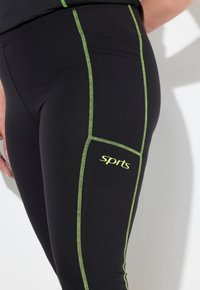 Leggings deportivos negros con costuras en verde neón contrastantes, que cuentan con un bolsillo lateral y la palabra "Deportes" impresa en la pierna.