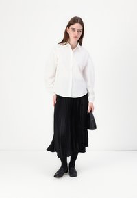 Biela košeľa s dlhými rukávmi a gombíkmi vpredu v kombinácii s čiernou plisovanou midi sukňou. Modelka drží malú čiernu kabelku. Čierne topánky a pančuchy.