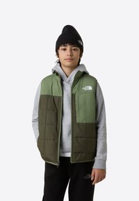 Vadderad väst i grön och oliv med dragkedja, har en framficka och logotyp. Bärs över en grå hoodie och svarta byxor.