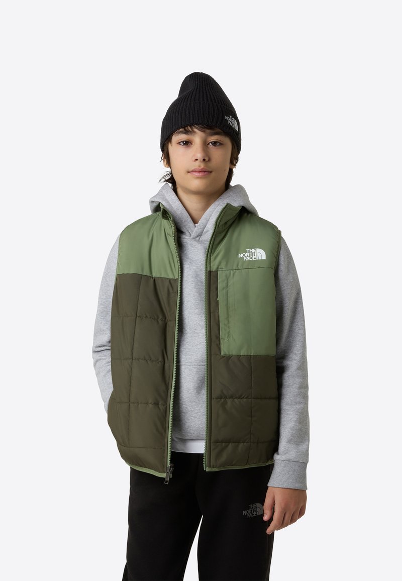 Vadderad väst i grön och oliv med dragkedja, har en framficka och logotyp. Bärs över en grå hoodie och svarta byxor.