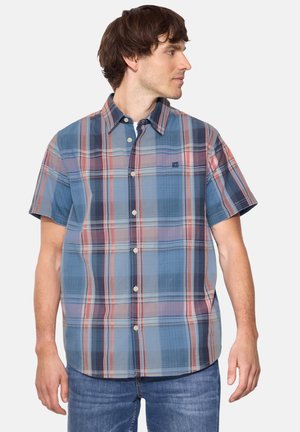 Mann trägt ein blau-rotes und graues kariertes Kurzarmhemd mit Knöpfen und blaue Jeans, schaut nach rechts vor weißem Hintergrund.