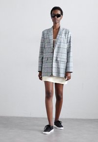 Grijze geruite oversized blazer met voorkhhouders, gecombineerd met een licht crèmekleurige jurk en zwarte sneakers. Draagster heeft moderne zonnebril op.