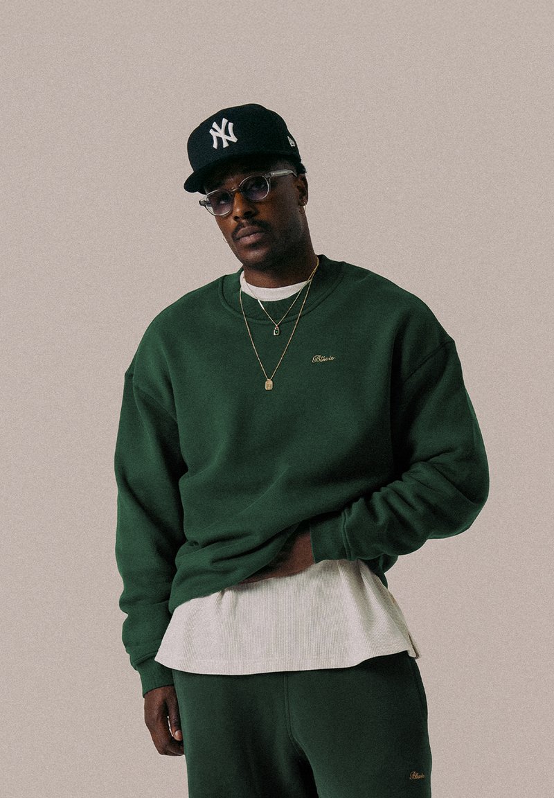 Grüner Fleece-Sweatshirt mit gerippten Bündchen, über einem weißen strukturierten Shirt getragen. Mit goldfarbenem Logodetail auf der Brust.