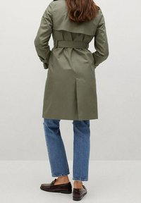 Personne portant un trench-coat ceinturé vert olive, un jean bleu et des mocassins brun foncé, debout de dos sur un fond uni.