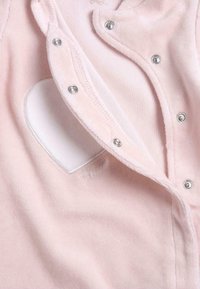 Ljusrosa bodysuit med tryckknappar, som har en vit hjärtformad utskärning och en mjuk struktur. Tillverkad av ett plyschmaterial med subtila sömnadsdetaljer.