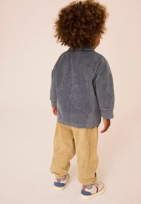 Harmaa corduroy-ylisuuri paita, jonka parina on vaaleanbeiget corduroy-housut. Lapsella on päällään valkoiset kengät, joissa on sinisiä ja vaaleanpunaisia yksityiskohtia. Kankaan tekstuuri näkyy.