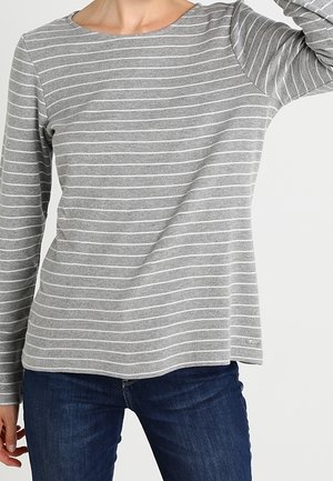 Frau trägt ein graues Langarmshirt mit weißen horizontalen Streifen und blaue Jeans vor einem schlichten hellen Hintergrund.