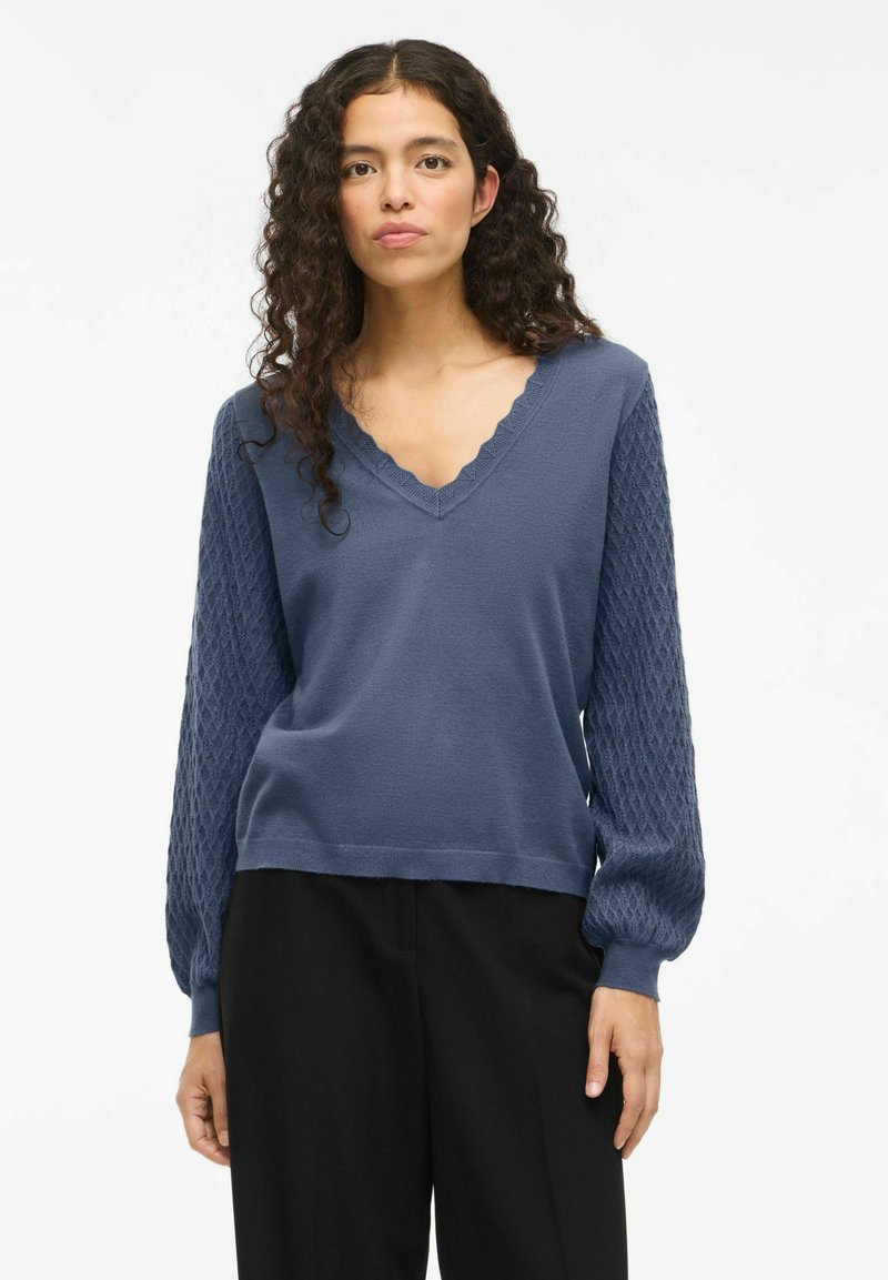 Suéter azul claro con un escote en V con puntas y mangas abullonadas texturizadas. Material suave con un cuerpo liso y un diseño de manga estampada.