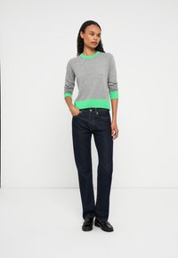 Jovem mulher está de pé com uma mão no bolso, vestindo um suéter cinza com detalhes em verde, jeans escuros e mocassins pretos, contra um fundo branco liso.