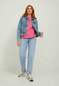 Jaqueta de ganga azul clara com fechos de botão, usada sobre um sweatshirt rosa. Jeans azuis claros e sapatilhas cinzas completam o visual.