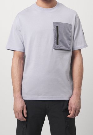 T-shirt gris à manches courtes en coton, avec une grande poche grise zippée de noir sur le côté gauche de la poitrine. Texture lisse.