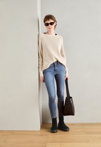 Pull côtelé beige, jeans ajustés bleus, bottines à plateforme noires, tenant un sac fourre-tout noir, debout contre un mur gris.