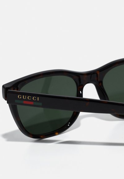 Ochelari de soare Gucci cu rame din material tartan, lentile verzi închise și logo auriu pe lateral. Au o formă unghiulară și o textură lucioasă.