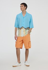 Camicia button-up azzurra a maniche corte con una grafica colorata in fondo. Pantaloni cargo arancioni con tasche laterali e vestibilità comoda. Sneakers bianche.