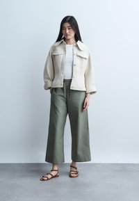 Mladá žena stojaca v krémovej bunde, bielom topu, olivových širokých nohaviciach a hnedých sandáloch s remienkami na jednoduchom svetlom pozadí.