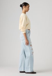 Femeie cu păr închis la culoare prins într-un coc, purtând un sweatshirt scurt de culoare bej deschis, blugi evazați, rupți, de culoare albastru deschis, și pantofi închiși la culoare, stând în profil.