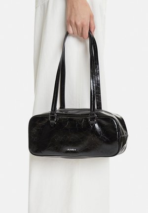 Hand hält eine kleine glänzende schwarze rechteckige Handtasche mit doppeltem Riemen und Reißverschluss, getragen über weißer Kleidung.