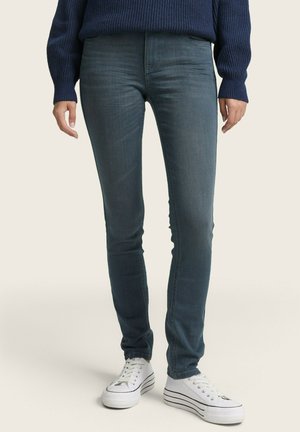 Dark blue skinny jeans with a smooth texture, slight stretch, and back pockets, paired with white sneakers featuring black stripes.